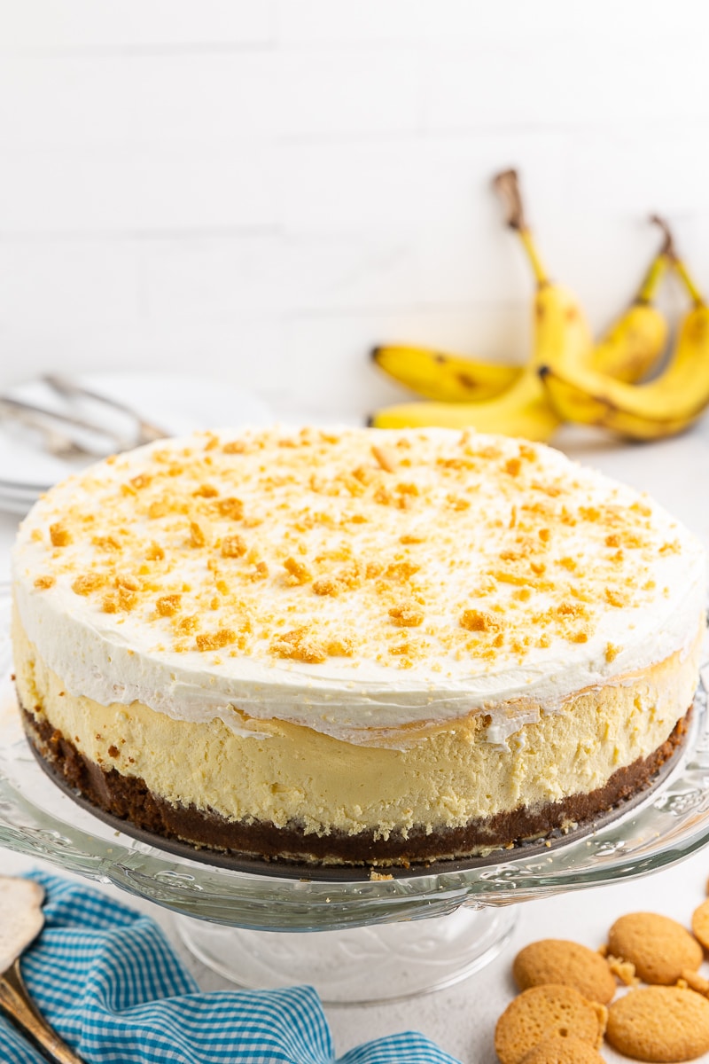 Keto Banana Pudding Cheesecake served on a glass cake stand, placed on a white marble counter. A stack of white plates and silver forks is positioned behind the cheesecake. Fresh bananas add a touch of vibrant color to the scene.