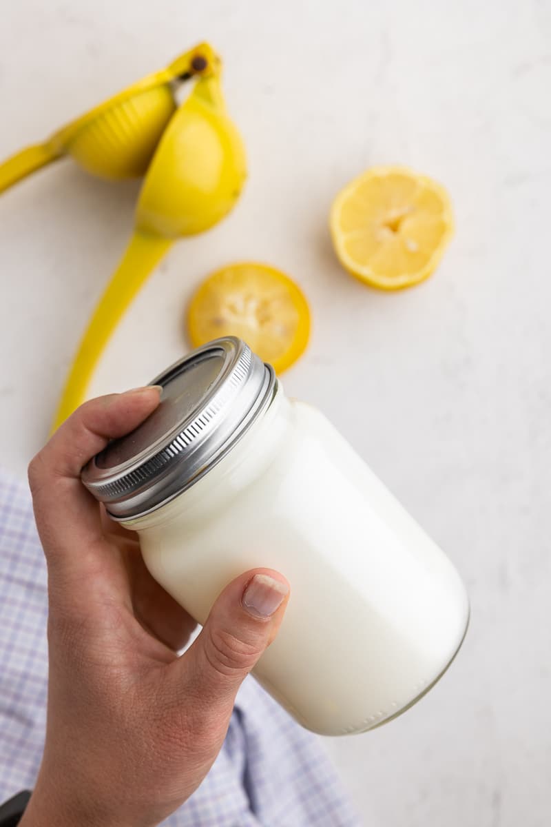 Hand holding mason jar with heavy cream and lemon juice for Keto KFC Coleslaw. White marble counter, blue and white towel, citrus juicer, and freshly squeezed lemon. Zesty touch for the low-carb coleslaw recipe!