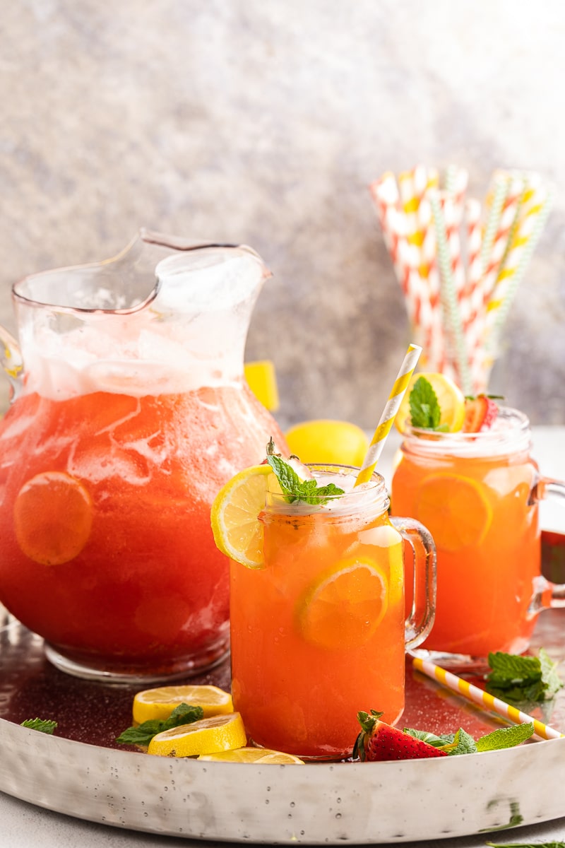 A glass pitcher full of Sugar-Free Strawberry Lemonade on a silver servings tray with 2 glass mugs full of lemonade. They have lemon slices, strawberry slices, and mint leaves as garnish and yellow and white paper straws.