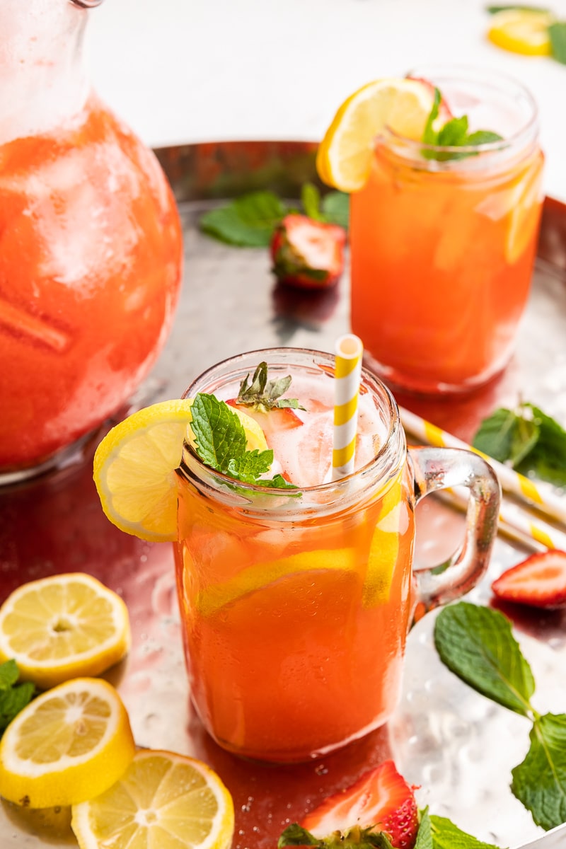 A glass mug full of Sugar-Free Strawberry Lemonade. The lemonade has slices of fresh lemon in the glass and is garnished with sliced lemon, sliced strawberry, mint leaves, and a bright yellow and white striped paper straw.