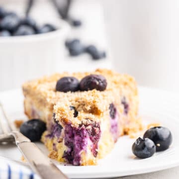 Close-up view of a tantalizing slice of Keto Blueberry Cream Cheese Coffee Cake on a pristine white plate, accompanied by a shiny silver fork. The slice showcases the luscious cream cheese filling inside, with burst blueberries creating a mesmerizing purplish-blue stain in the creamy filling. In the background, a slightly blurred cake stand holds the remaining coffee cake, while a bowl of blueberries and another plate with a slice of cake add to the display. A white mug of coffee completes the scene, providing a cozy backdrop.