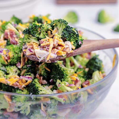 Close up side view of a glass bowl full of Loaded Broccoli Salad. You can see the brilliant green of the broccoli, red of the onion, orange from the cheese. It is very visually enticing.