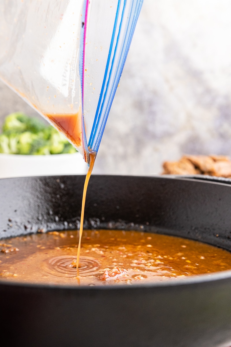 Side view of pouring leftover marinade into the skillet to create the flavorful sauce for the Keto Korean Beef. A zip lock baggie is held above the cast iron skillet, allowing the marinade to flow gracefully into the pan. Broccoli and cooked beef are slightly blurred in the background, adding to the culinary scene.