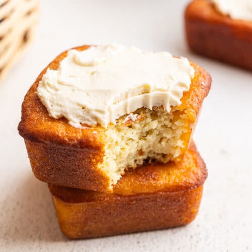 Two keto cornbread muffins stacked on a clean white counter. The upper muffin is adorned with a dollop of sugar-free maple butter, and a single bite has been taken out of it, revealing its moist interior. The golden-brown crust of the muffins hints at their satisfying texture. The image captures the deliciousness of a guilt-free treat.