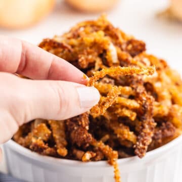 Close-up view of Keto Fried Onions in a white ramekin, with a hand holding one onion closer to the camera. The crispy low carb onions add a delightful crunch to dishes.