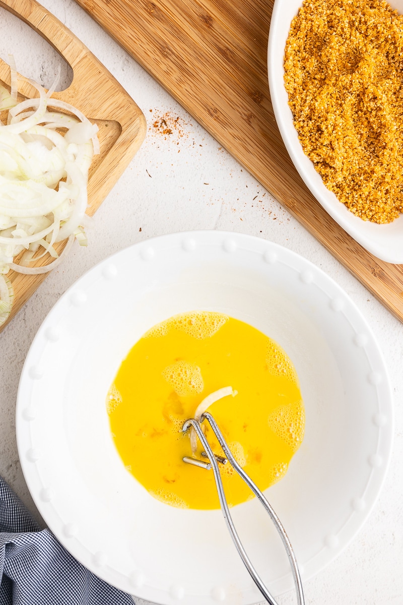 From above, a white bowl holds beaten eggs, with a slice of onion gripped by metal tongs immersed within. Adjacent to the eggs, sliced onions rest on a wooden cutting board to the upper left. Nearby, a shallow white dish contains the ground pork rind mixture, while a blue and white kitchen towel is positioned to the bottom left.
