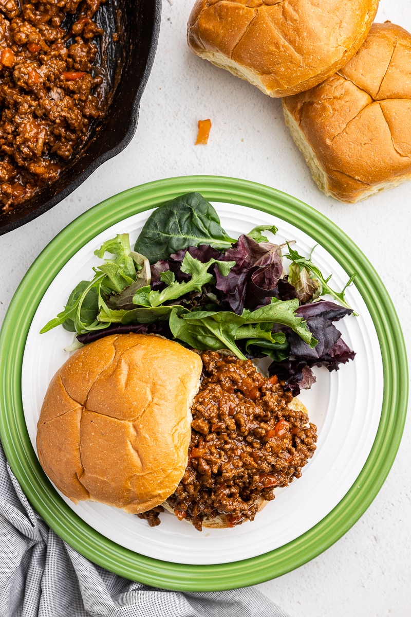 Delicious Keto Sloppy Joes Meal - Overhead view of the keto sloppy joe on a white plate with a green rim, accompanied by a side of mixed greens salad. The plate is placed on a white counter. In the upper left corner, the cast iron skillet contains the remaining sloppy joe mixture, and the upper right corner features two keto buns. A satisfying and vibrant low-carb meal presentation.
