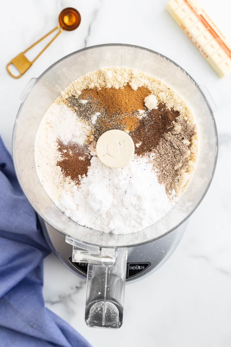 Image shows almond flour, coconut flour, powdered sweetener, baking powder, cinnamon, ginger, cardamom, allspice, cloves, pepper, and xanthan gum in a food processor on a white counter.