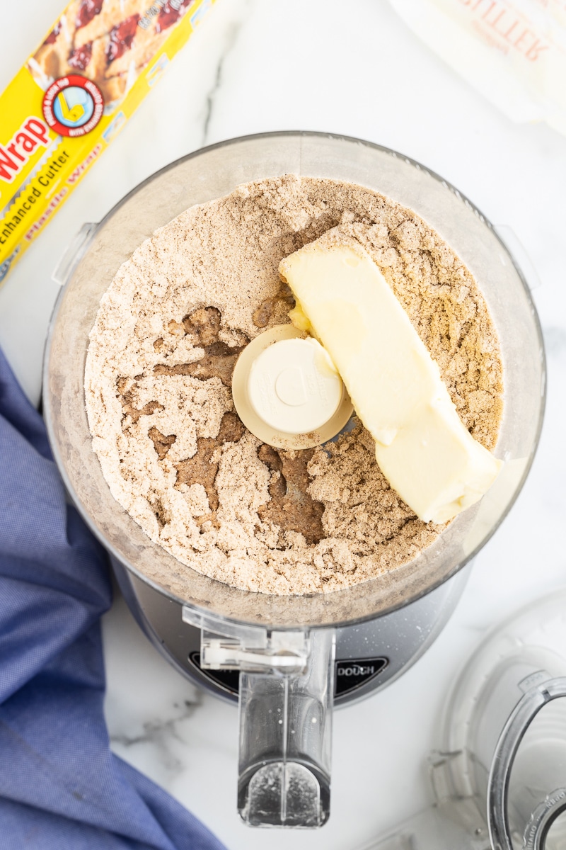 Image shows a stick of softened butter being added to the bowl of a food processor that contains almond flour, coconut flour, powdered sweetener, baking powder, cinnamon, ginger, cardamom, allspice, cloves, pepper, and xanthan gum.