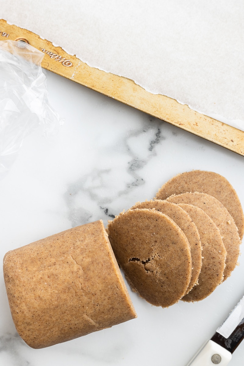 Image shows keto chai sugar cookie dough being sliced into discs for baking on a white marble counter.