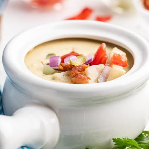 Image shows a small white crock with a handle full of Keto Cheeseburger Soup topped with crispy bacon and chopped tomato, red onion, and pickles.