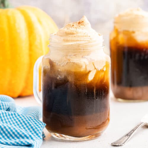 The featured image shows a mason jar mug with keto pumpkin cold brew topped with sugar-free pumpkin cold foam on a white counter with a silver spoon to the right of the mug. In the background there is another mug of the pumpkin cold brew to the right and a pumpkin to the left.