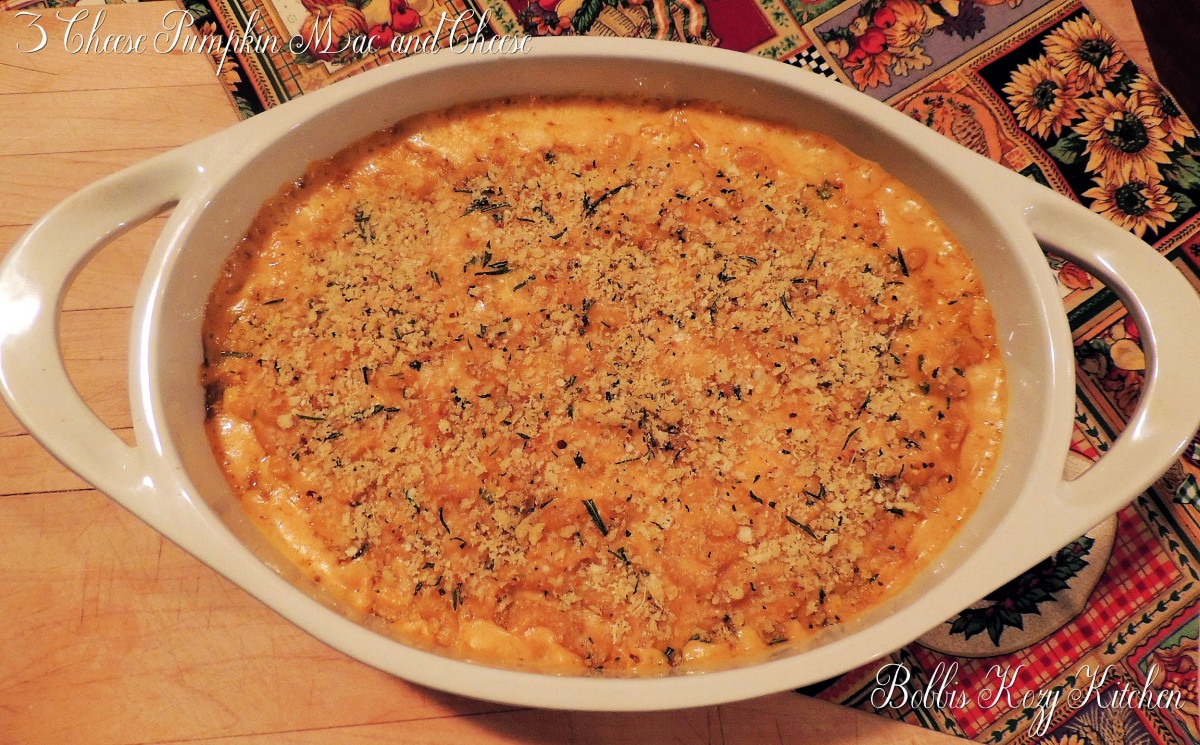 Overhead view of a white oval casserole dish with 3 cheese pumpkin mac and cheese topped with toasted keto bread crumbs in it. The casserole dish is sitting on a wooden cutting board with an fall themed place may under the casserole dish.