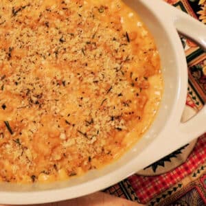 Closeup of the corner of a white casserole dish full of 3 cheese pumpkin mac and cheese topped with keto brad crumbs on a wooden cutting board with a autumn themed placemat under the casserole dish.