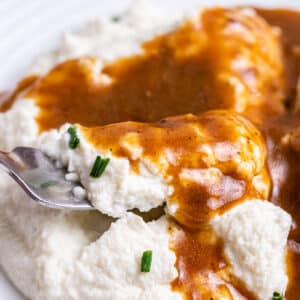 Closeup of mashed cauliflower on a white plate smothered in keto gravy with a silver fork getting a bite.
