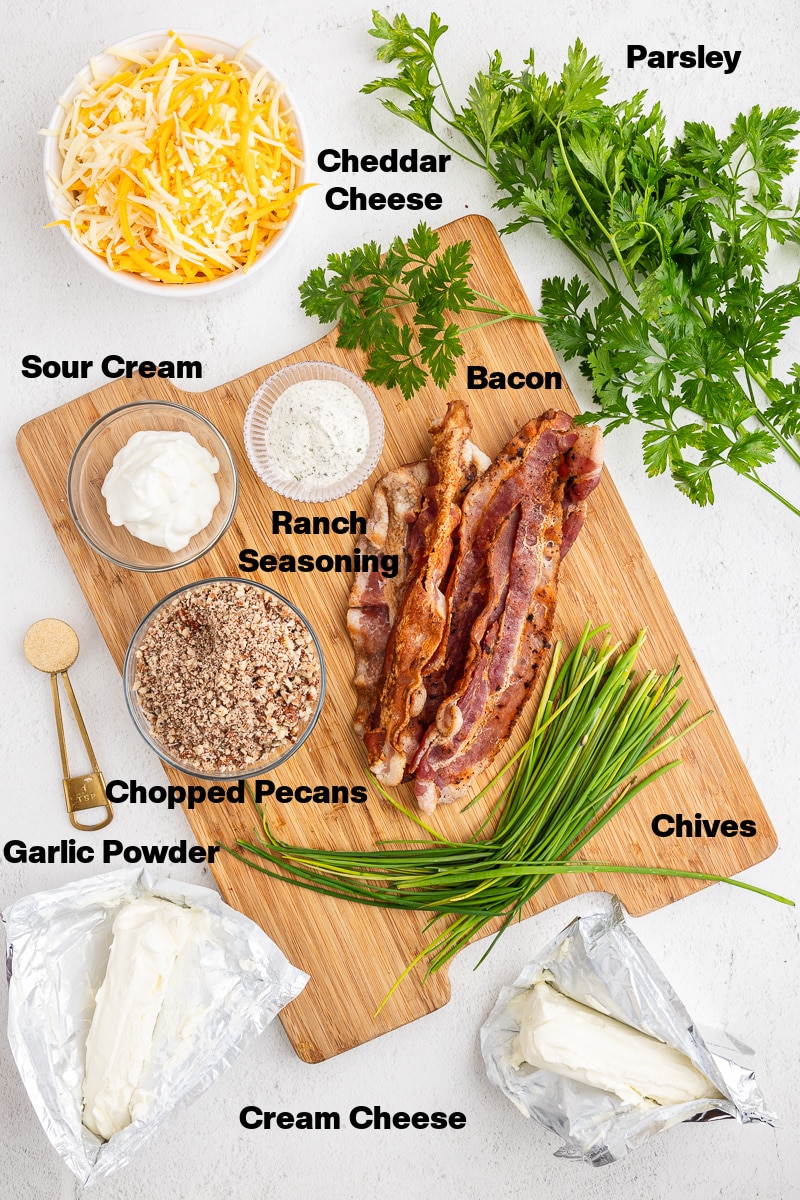 Overhead view of all of the ingredients needed to make a keto bacon ranch cheese ball on a wooden cutting board and a white counter.