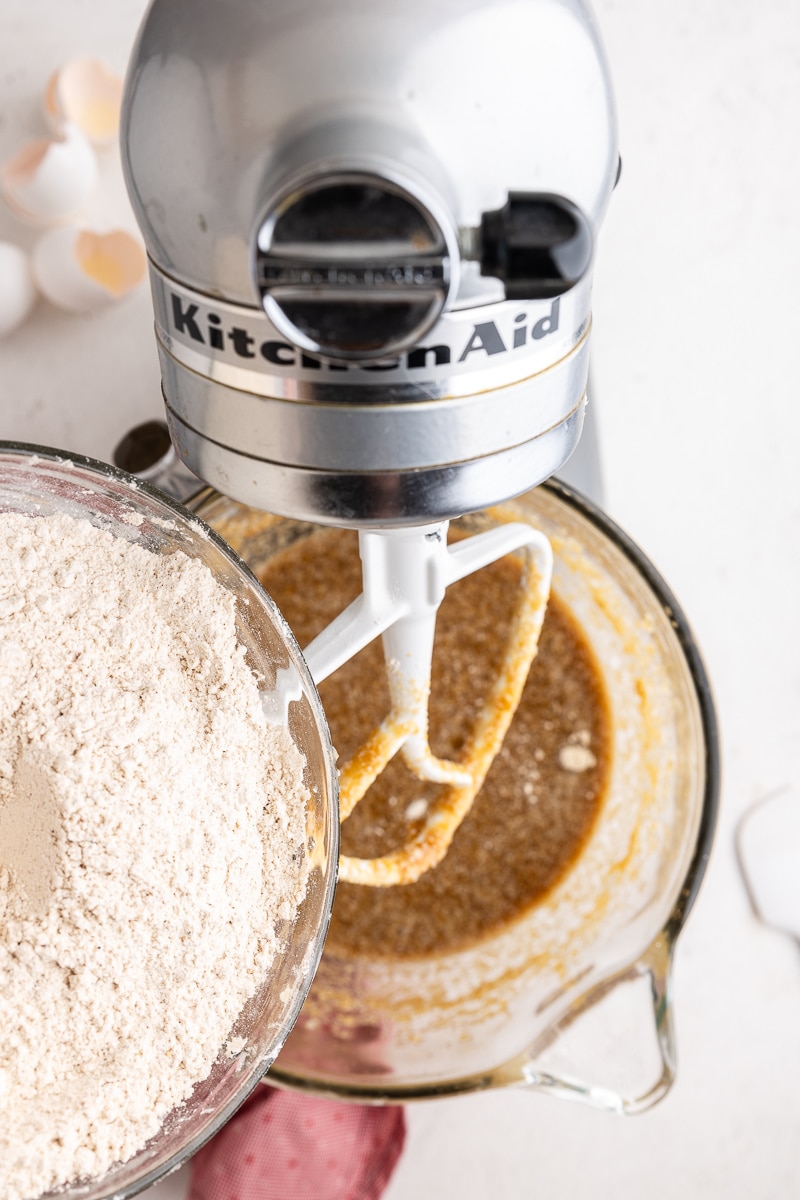 Overhead view of the wet ingredients for the keto Christmas spice cake in the bowl of a stand mixer with a large bowl full of the dry ingredients being poured in.