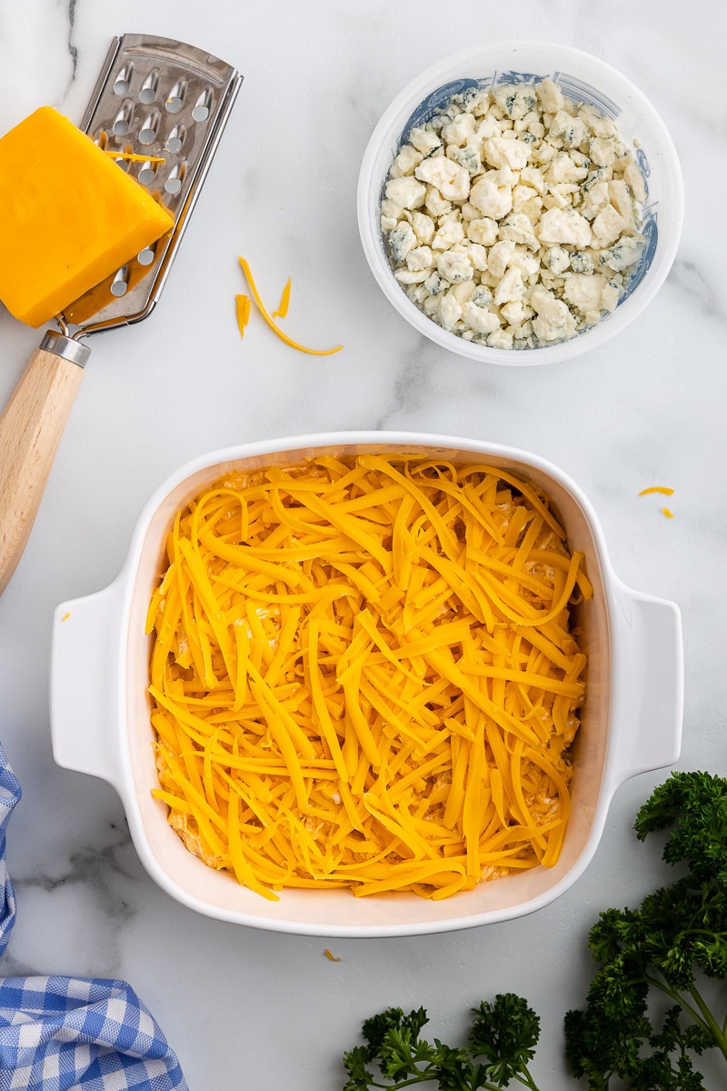 Overhead view of a white casserole dish full of Buffalo chicken dip and topped with shredded cheddar cheese right before it goes into the oven.