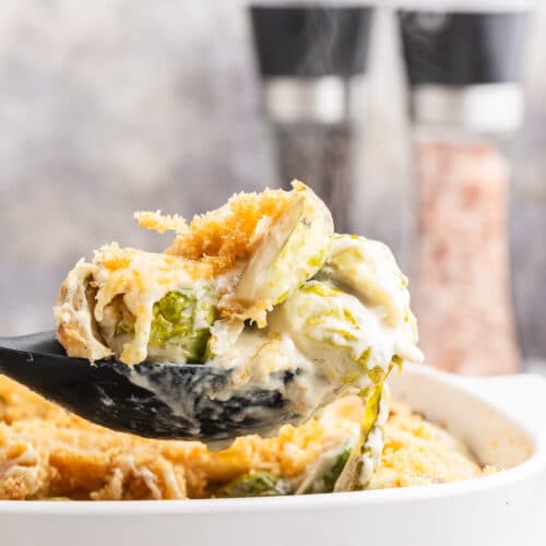 Keto brussels sprouts gratin in a white casserole dish with a black serving spoon holding a spoonful over the dish.
