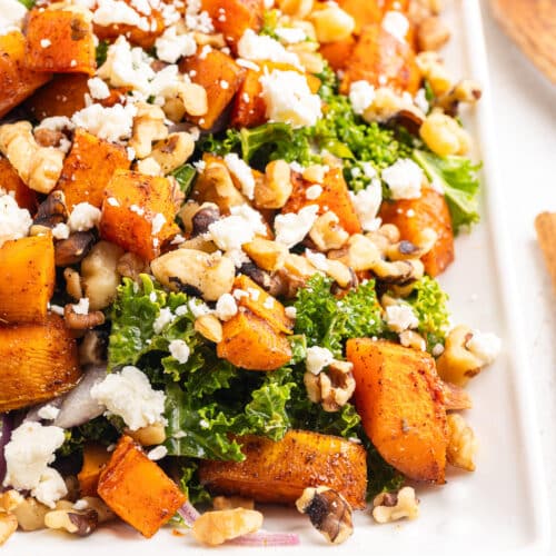 Closeup of a Roasted Butternut Squash and Kale Salad with goat cheese crumbles and toasted walnuts on a white serving platter.