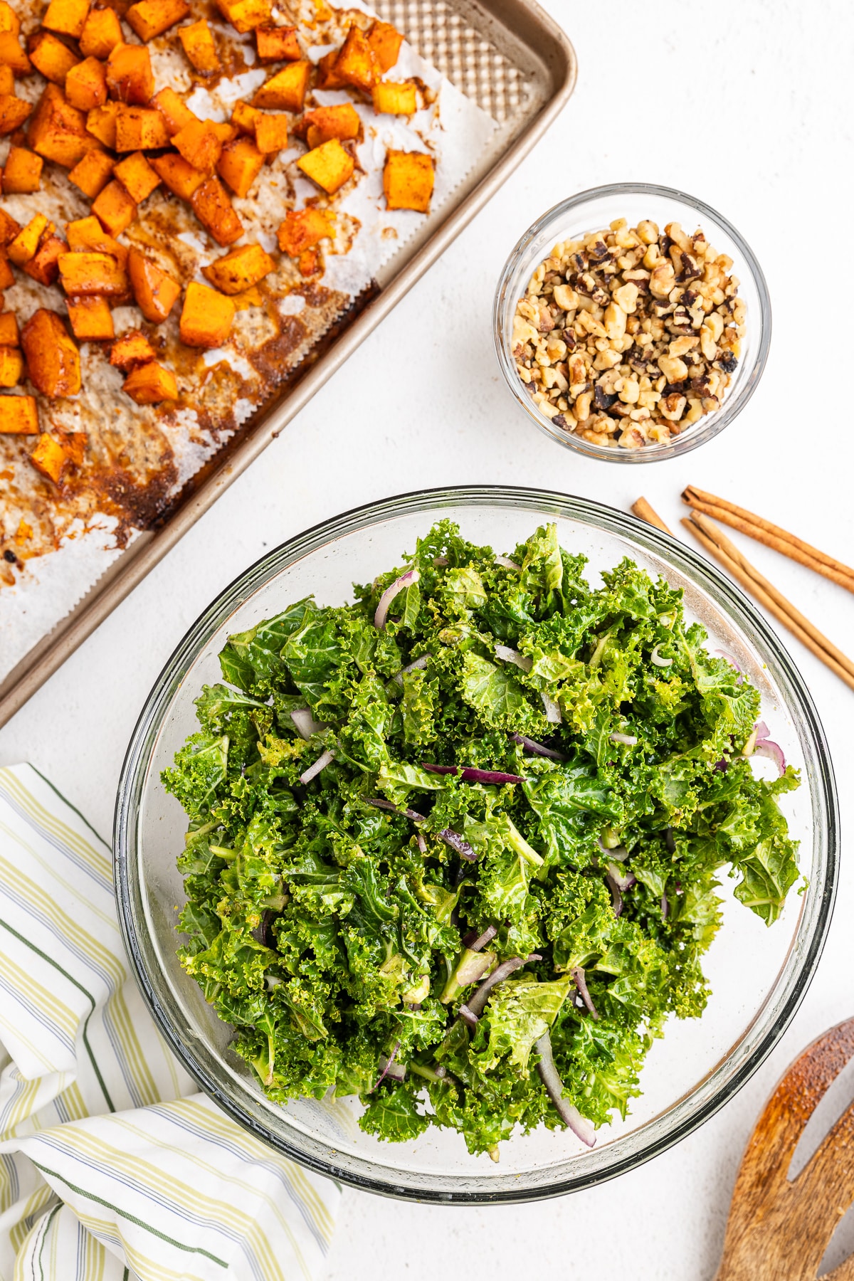 Overhead view of a large glass bowl full of baby kale and red onion tossed with an orange chili salad dressing next to a baking sheet full of roasted butternut squash cubes. There is also a small glass bowl full of chopped walnuts.
