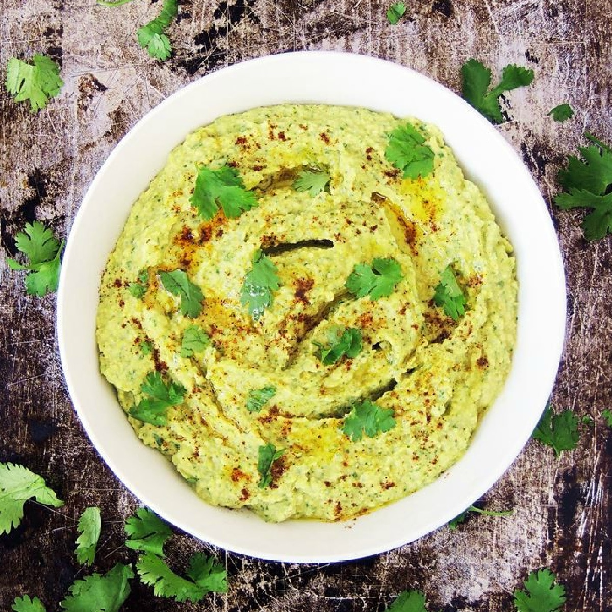 Overhead view of a bowl of roasted jalapeno hummus on a metal baking sheet with fresh cilantro leaves scattered about.