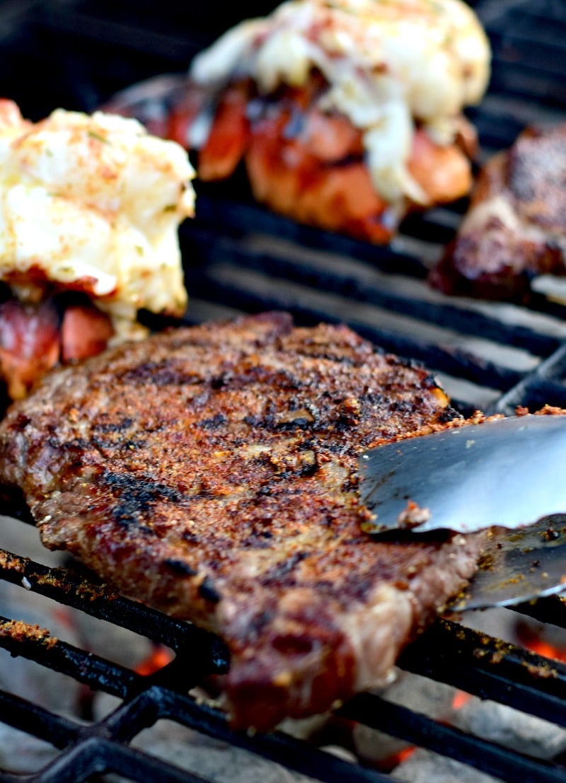 A perfectly grilled ribeye being held with silver tongs over a grill. In the background you can see lobster tails on the grill.