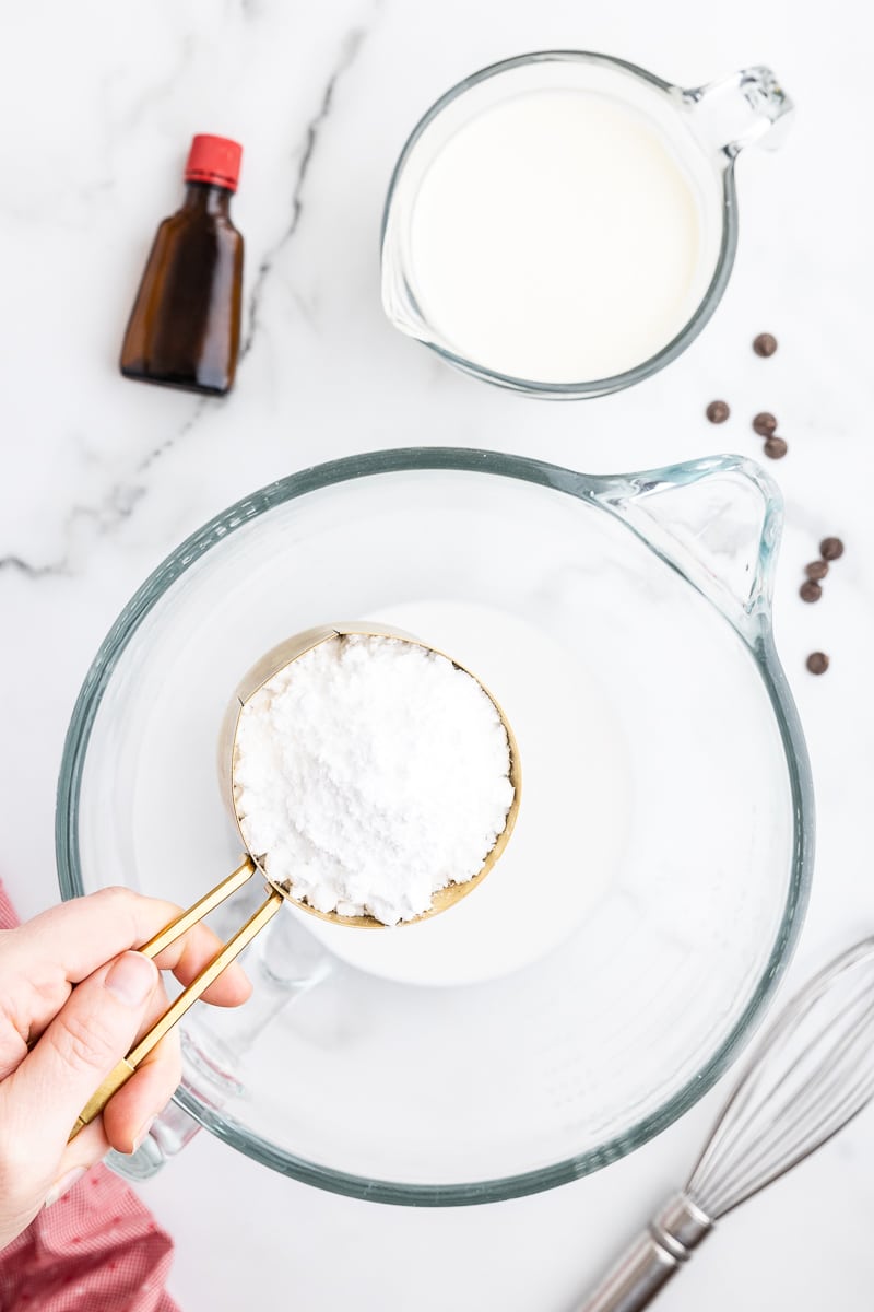 Overhead view of the glass mixing bowl for a stand mixer with coconut milk in it and a white female presenting hand holding a gold measuring cup full of keto powdered sweetener above it.