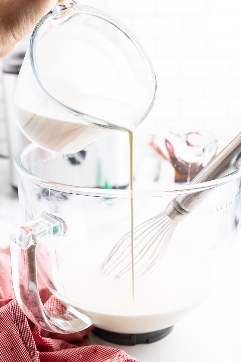 Heavy cream being poured from a glass measuring cup into the bowl of a stand mixer containing coconut milk and keto powdered sweetener.