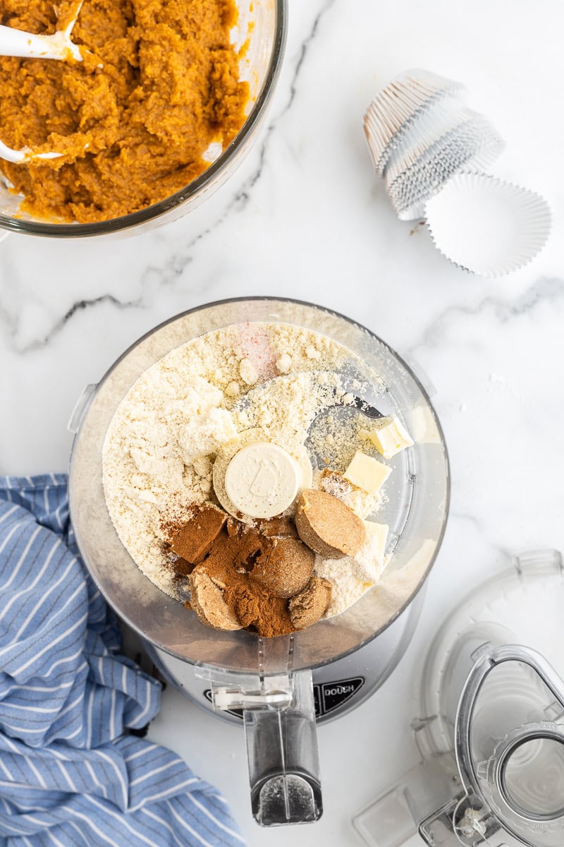 Overhead view of the ingredients for the streusel topping in the bowl of a food processor, ready to be combined. To the top left, a bowl contains the pumpkin muffin mixture, showcasing the preparation process for Keto Pumpkin Cream Cheese Muffins.