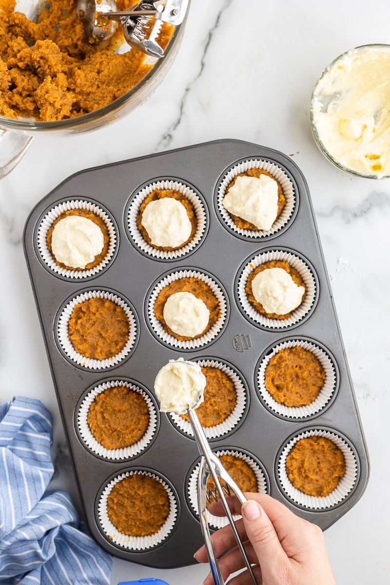 Overhead view of a standard muffin pan lined with muffin liners filled with pumpkin muffin batter. A white female-presenting hand is scooping small amounts of cream cheese mixture into each muffin cup, preparing for the final baking of Keto Pumpkin Cream Cheese Muffins.