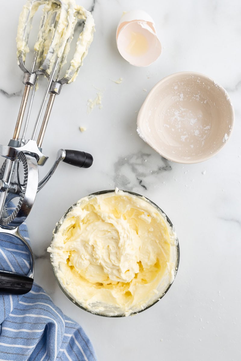 Overhead view of a small glass mixing bowl filled with the combined ingredients for the cream cheese filling. The bowl is placed on a white counter alongside a set of manual hand beaters with cream cheese filling on them, and a couple of empty small bowls that previously contained some of the filling ingredients.