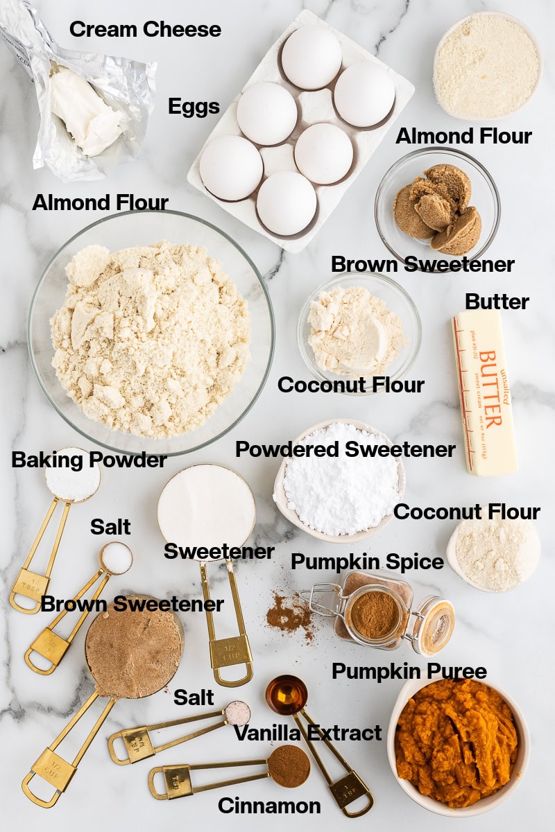 Overhead view of all of the ingredients needed to make these keto pumpkin cream cheese muffins on a white marble background. Each ingredient is labeled.