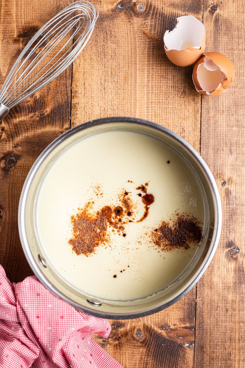 Overhead view of a silver sauce pot containing coconut milk, eggs, keto sweetener, and salt. There is vanilla extract and nutmeg sitting on the surface before being combined into the keto eggnog base.