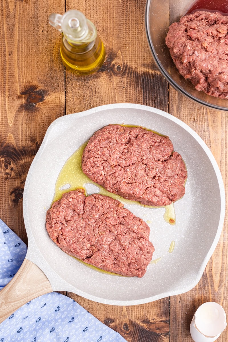 Overhead view of two keto salisbury steak patties in a white ceramic skillet on a wooden table.