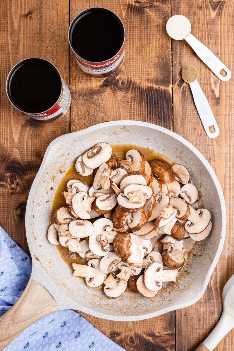 Overhead view of sliced brown mushrooms sauteing in a white cermaic skillet to make the mushroom gravy.