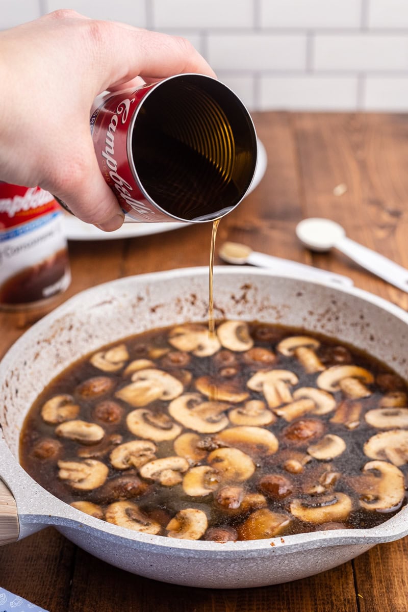 Closeup of a white hand pouring beef consomme into the skillet to make mushroom gravy.