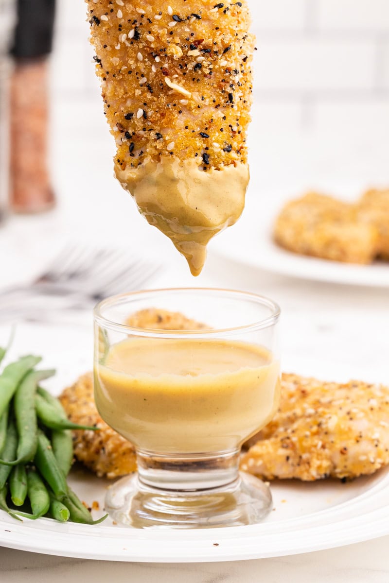 Closeup of an everything bagel chicken tender being dipped into a small glass bowl containing sugar-free maple mustard.