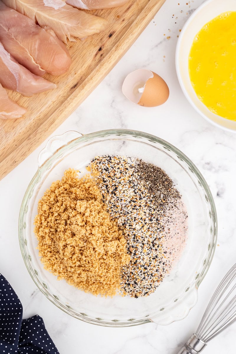 Overhead view of a glass bowl containing ground pork rinds, everything bagel seasoning, salt and pepper.