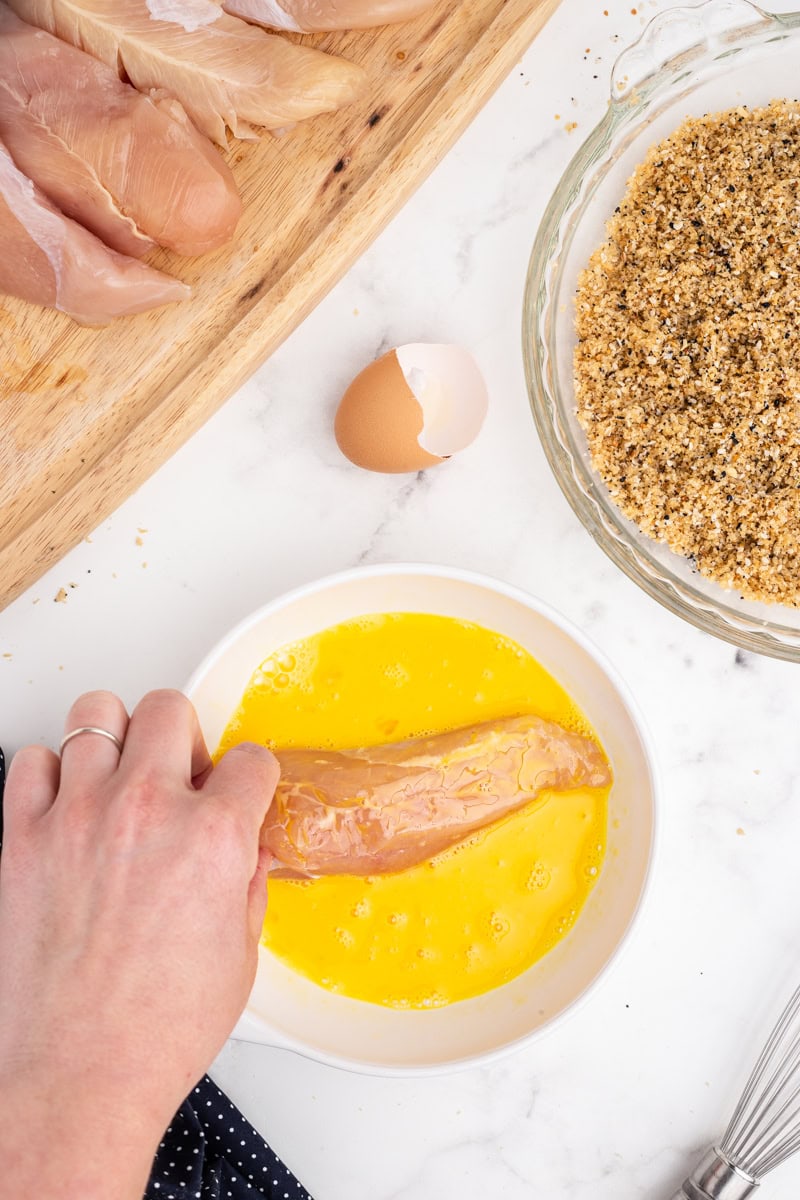 Overhead view of a white female presenting hand dipping a chicken tender into a white bowl containing beaten egg.