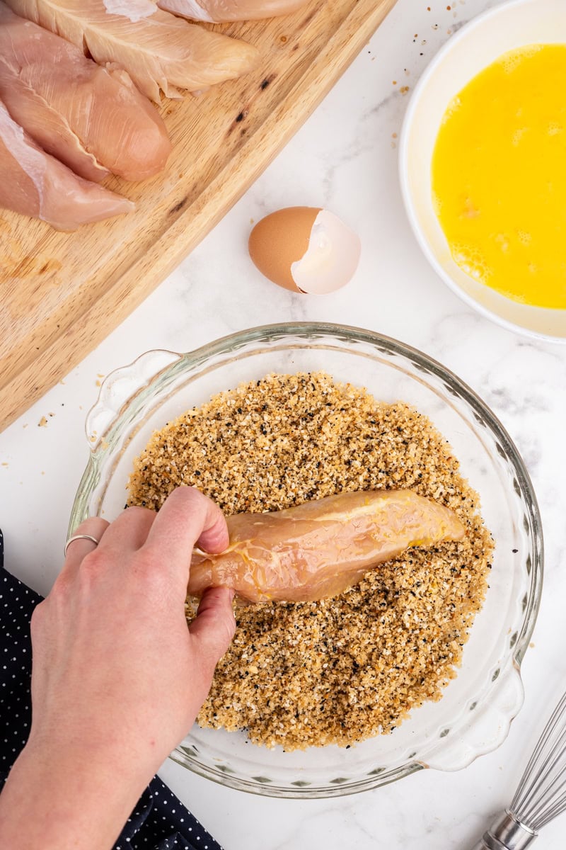 Overhead view of a white female presenting hand dredging a chicken tender coated in beaten egg into a mixture of everything bagel seasoning, ground pork rinds, salt, and pepper.