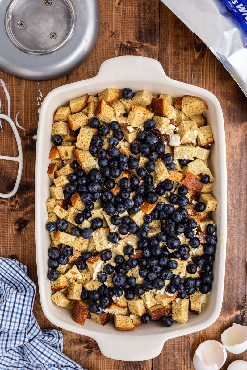Keto bread cubes and custard mixture topped with blueberries in a 9 x 13 baking dish on a wooden table.
