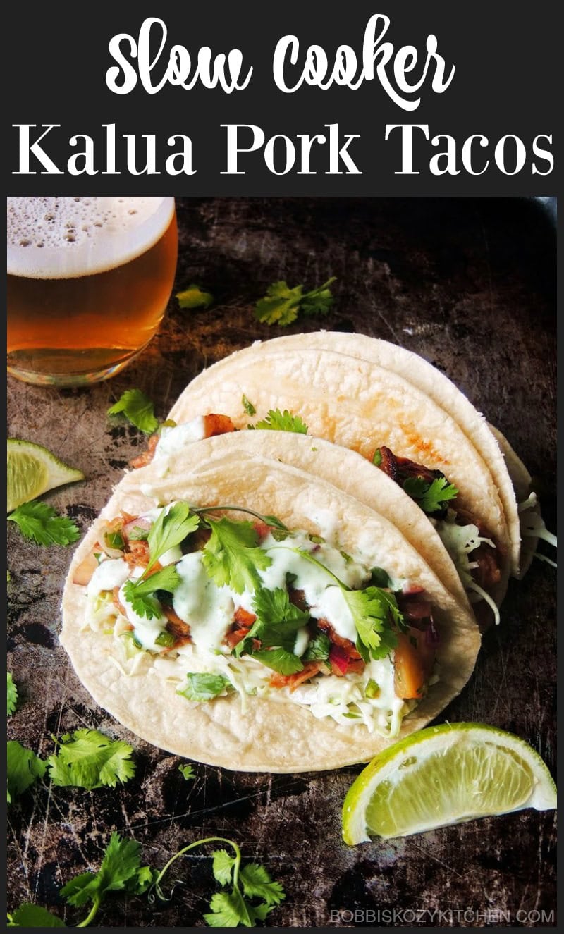 Three sugar-free Slow Cooker Kalua Pork Tacos on a metal table with lime slices and chopped fresh cilantro scattered around them.