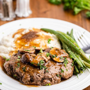 Keto Salisbury Steak with mushroom gravy on a white dinner plate with mashed cauliflower and steamed green beans.