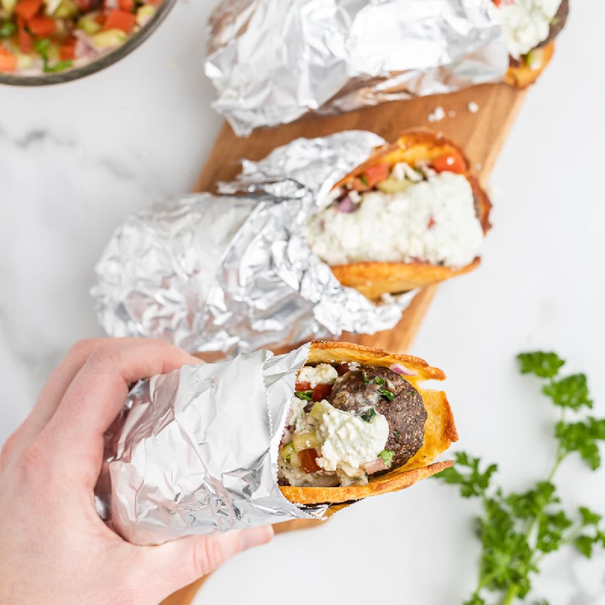 A white female presenting hand holding a low-carb gyro sandwich wrapped in foil with two more laying on a wooden cuttin gboard in the background.
