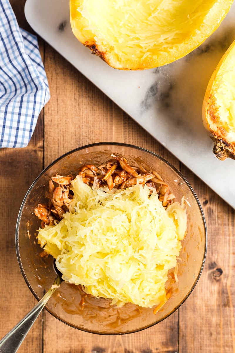Shredded chicken with barbeque sauce and spghetti squash strands in a glass bowl on a wooden table with two roasted spaghetti squash halves beside on on a marble cutting board.