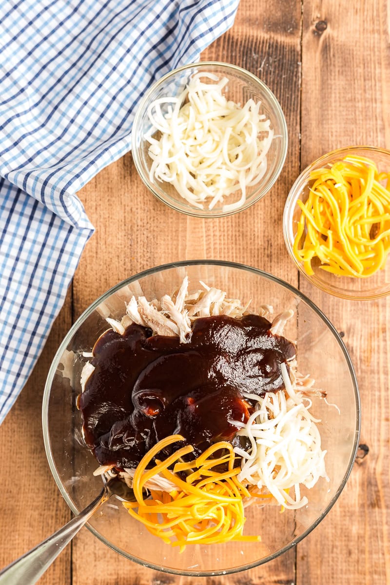 Shredded chicken, barbeque sauce, and shredded cheese in a large glass bowl on a wooden table.