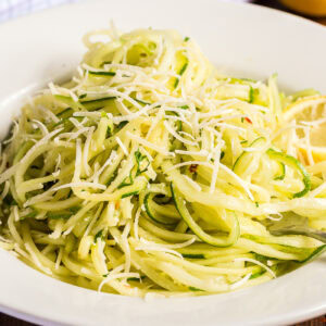 Closeup of a serving of lemon garlic zucchini noodles in a white bowl. The zoodles are topped with fresh grated parmesan and chopped parsley.