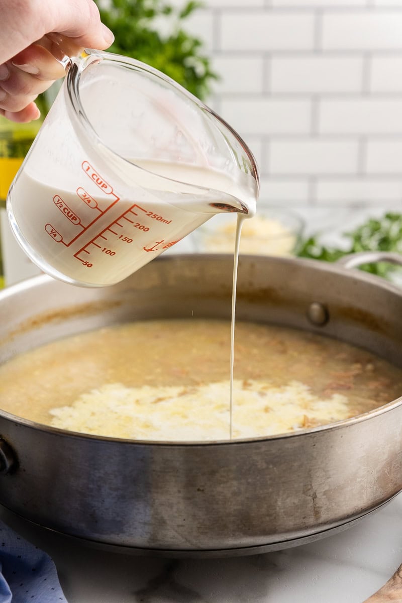 A white female presenting hand pouring heavy cream into a large skillet containing shallots, garlic, baby clams, and white wine.
