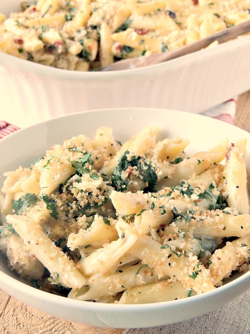 Closeup of a white bowl full of Chicken Florentine Casserole on a wooden table. You can see the baking dish containing the remaining casserole in the background.
